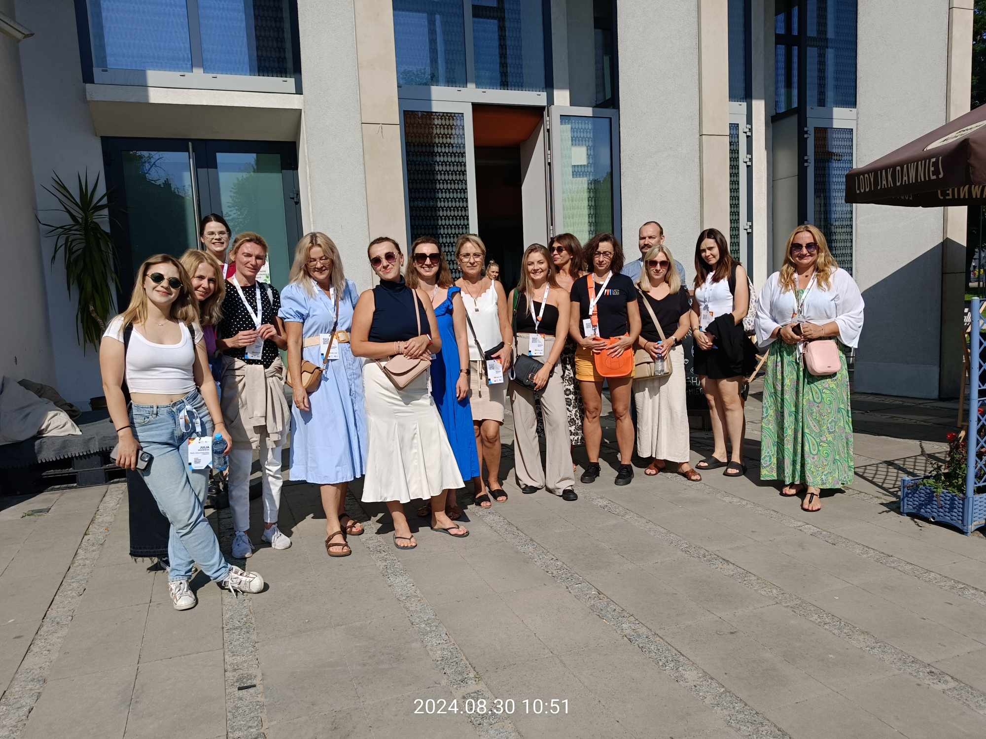 zdjęcie przedstawia uczestników tegorocznej edycji projektu ,,Zaproś nas do siebie", którzy odwiedzili MDK Ratusz 30 sierpnia w ramach wizyty studyjnej. Kilkanaście osób stoi na tle budynku Ratuszu od strony wejścia do kina.