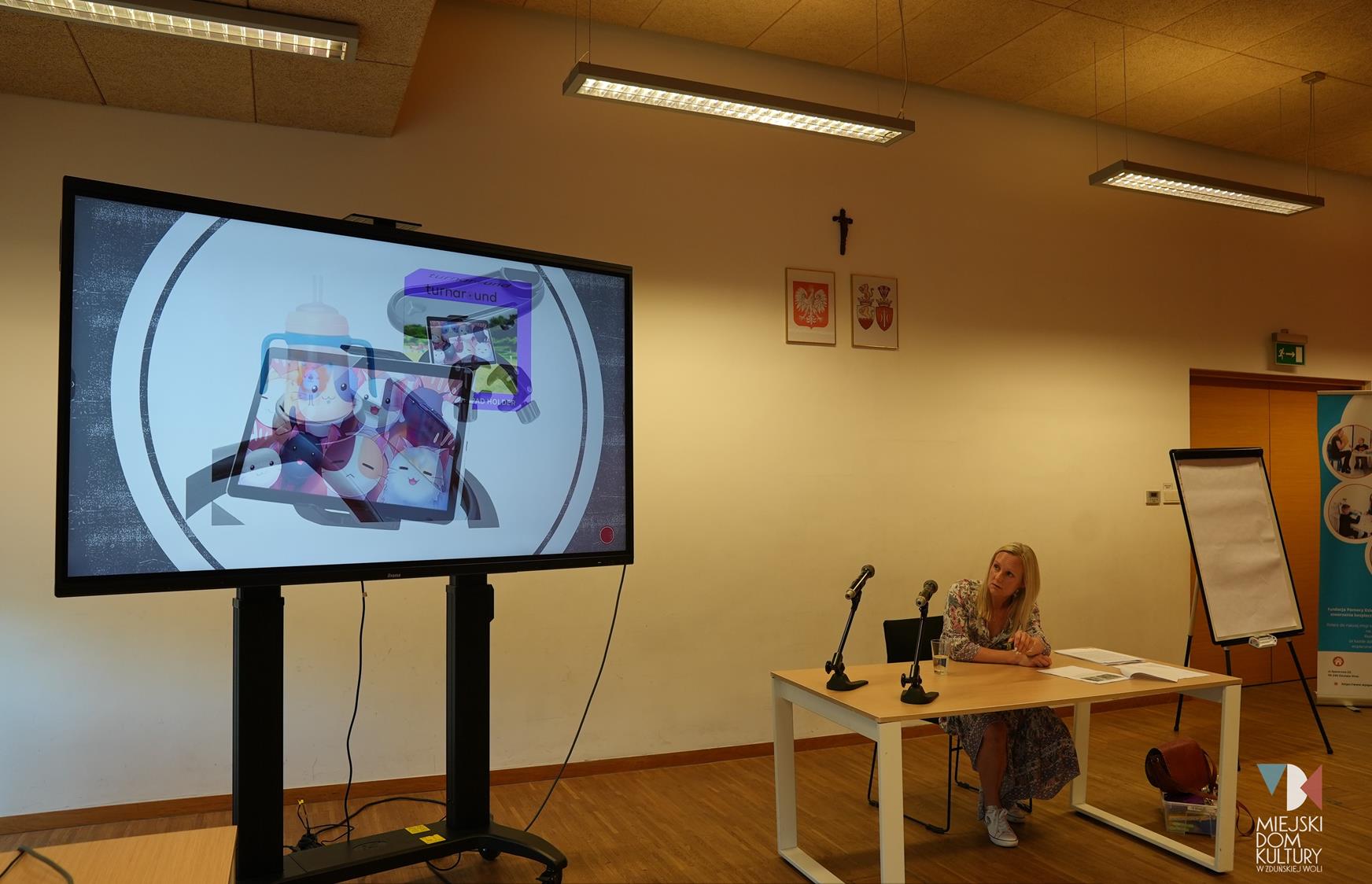 zdjęcie przedstawia na tablicę multimedialną i prelegentkę, kobietę w długich włosach blond, która siedzi przy stole i opowiada o dorastaniu w sieci