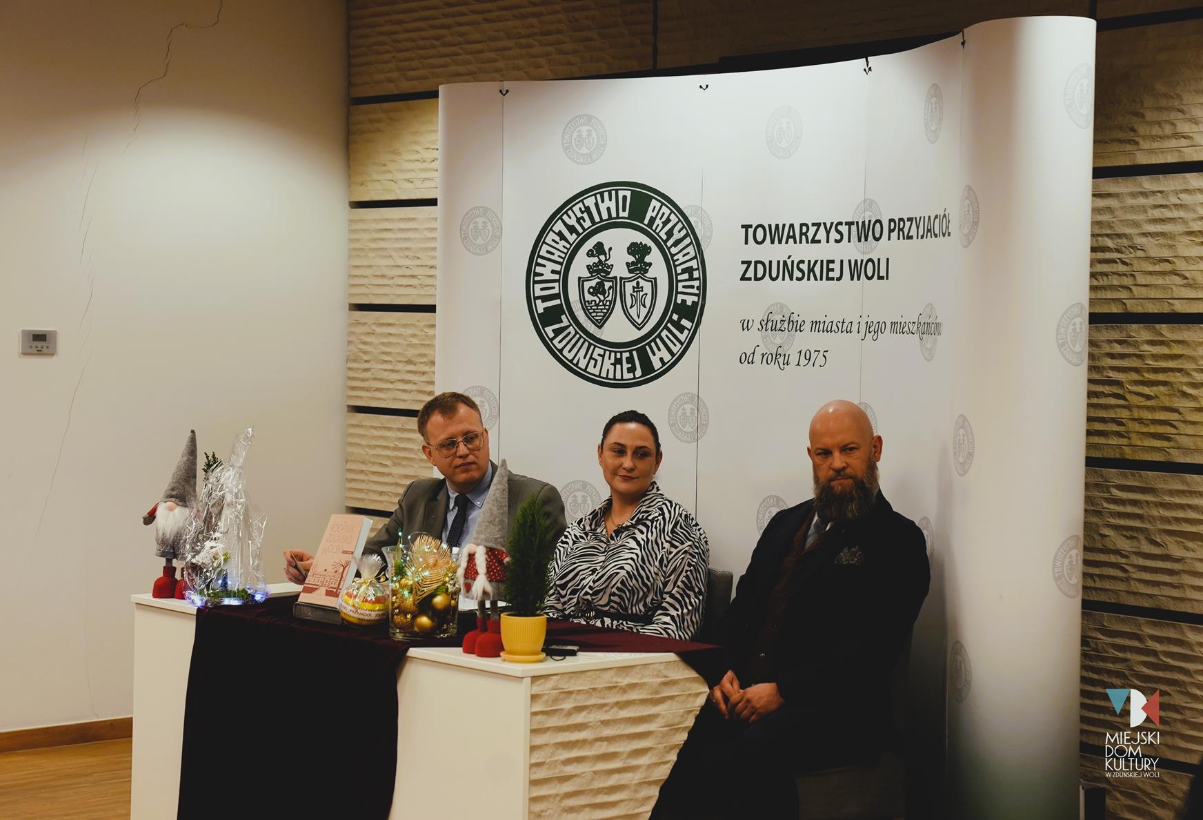 zdjęcie przedstawia trzy osoby, kobietę w czarnych włosach i biało-czarnej sukience i dwóch młodych mężczyzn , jeden jest w okularach , drugi ma brodę, wszyscy siedzą przy stole