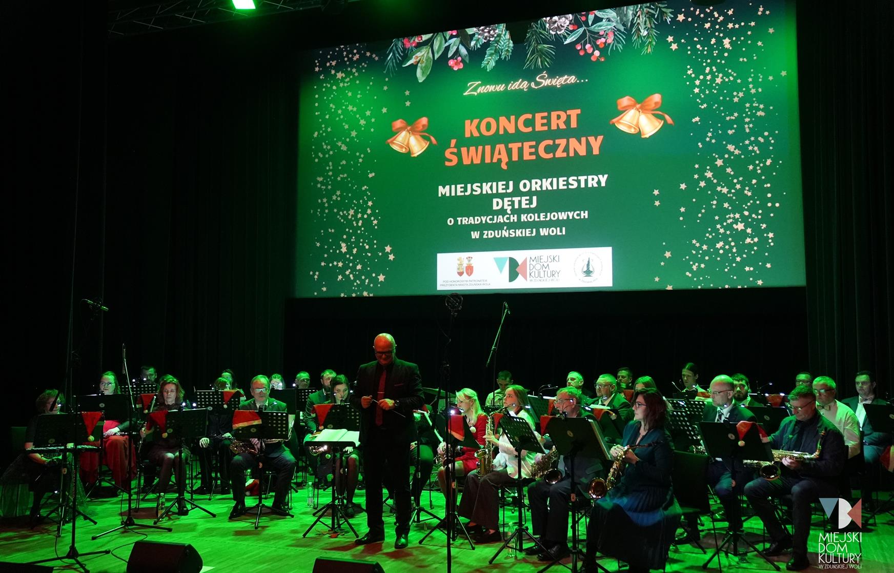 zdjęcie przedstawia ok. 40 osób , to muzycy Miejskiej Orkiestry Dętej o Tradycjach kolejowych. Muzycy występują w Ratuszu, stoją na scenie podczas Koncertu Świątecznego