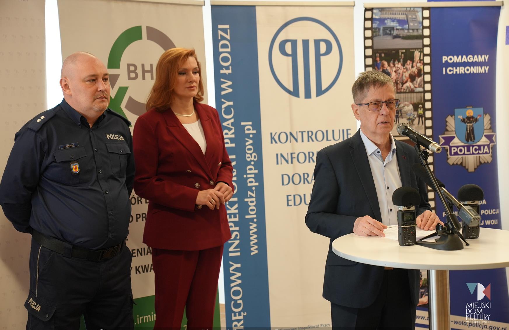 zdjęcie przedstawia trzy osoby, dwie kobiety i mężczyznę, którzy stoją na tle ścianki z rollupów, dwaj mężczyźni i kobieta zapraszają na konferencję dot. bhp, jaka odbędzie się w MDK Ratusz 9 kwietnia