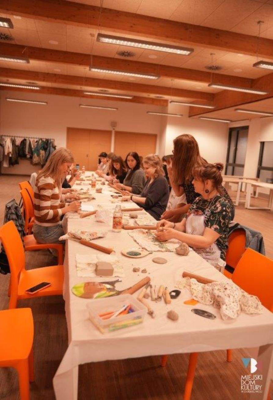 zdjęcie przedstawia uczestniczki warsztatów ceramicznych przy stole nakrytym białym obrusem, kobiety zajmują się lepieniem naczyń, podczas warsztatów ceramicznych w sali konferencyjnej MDK Ratusz