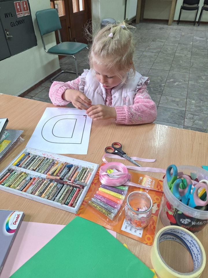 zdjęcie przedstawia małą dziewczynkę w jasnych włosach, która przygotowuje laurkę z kartki papieru na dzień mamy