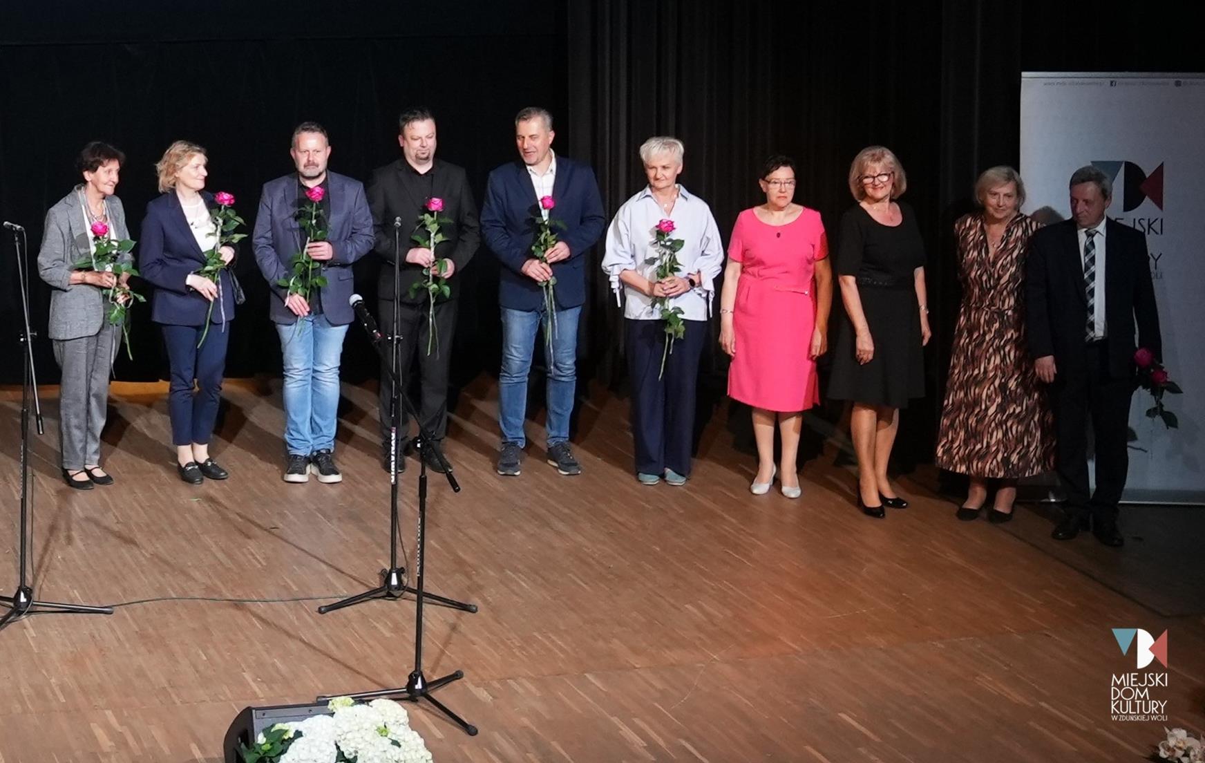zdjęcie przedstawia kilkanaście osób, 10 osób na scenie w Ratuszu z różami w dłoni, to instruktorzy i zarząd UTW