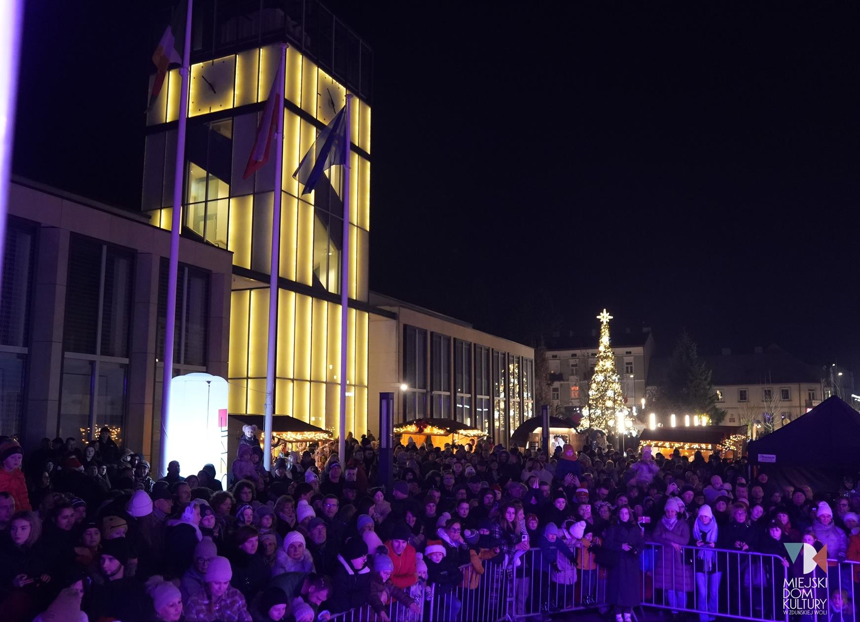 zdjęcie przedstawia budynek Ratusz oświetlony w tle mnóstwo ludzi, rozświetlona choinka. To wszystko na Jarmarku Świątecznym