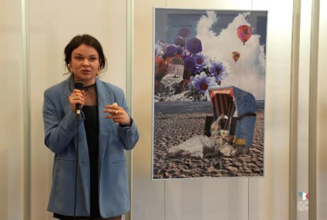 zdjęcie przedstawia młodą kobietę w ciemnych upiętych włosach w jasno niebieskim żakiecie z mikrofonem na tle kolorowej pracy z kolażem