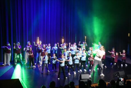 Zdjęcie przedstawia kilkanaście os&oacute;b w kolorowych strojach, to uczestnicy zajęć tanecznych MDK Ratusz na scenie w sali widowiskowej