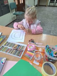 zdjęcie przedstawia małą dziewczynkę w jasnych włosach, która przygotowuje laurkę z kartki papieru na dzień mamy