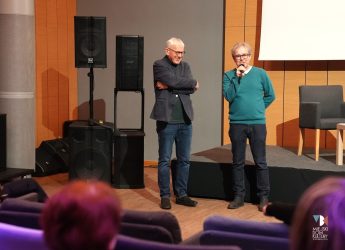 zdjęcie przedstawia dwóch mężczyzn w średnim wieku, jeden w zielonym swetrze z mikrofonem w dłoni i okularach, drugi w ciemnym ubraniu, lekko pochylony, obaj stoją w sali kameralnej MDK Ratusz