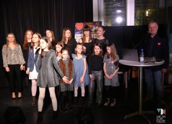 zdjęcie przedstawia kilkanaście osób, to młode wokalistki ze Studia Piosenki MDK Ratusz, obok stoi starszy mężczyzna w marynarce w krótkich włosach, to ich instruktor Andrzej Wawrzyniak