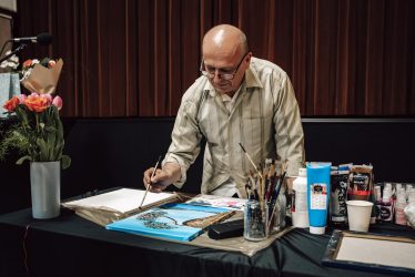 zdjęcie przedstawia starszego mężczyznę bez włosów pochylonego nad pracą plastyczną