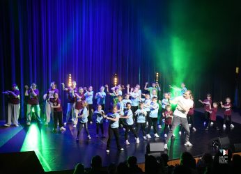 Zdjęcie przedstawia kilkanaście osób w kolorowych strojach, to uczestnicy zajęć tanecznych MDK Ratusz na scenie w sali widowiskowej